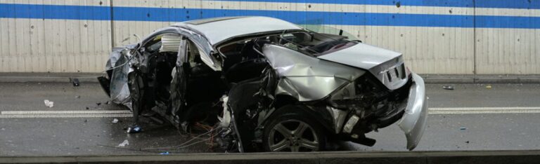 Stark zerstörtes silbernes Auto nach einem Unfall in einer tunnelartigen Strasse; das Fahrzeug ist an Front, Seite und Heck massiv beschädigt und steht auf nasser Fahrbahn vor einer Betonwand mit blauen Streifen.