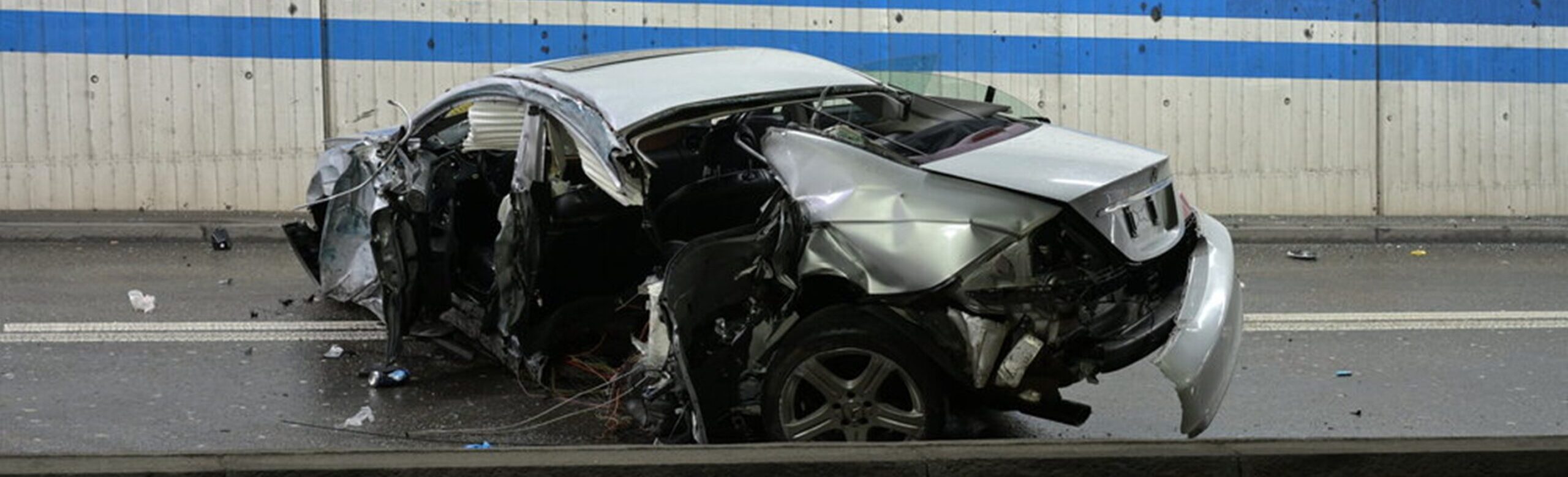 Stark zerstörtes silbernes Auto nach einem Unfall in einer tunnelartigen Strasse; das Fahrzeug ist an Front, Seite und Heck massiv beschädigt und steht auf nasser Fahrbahn vor einer Betonwand mit blauen Streifen.