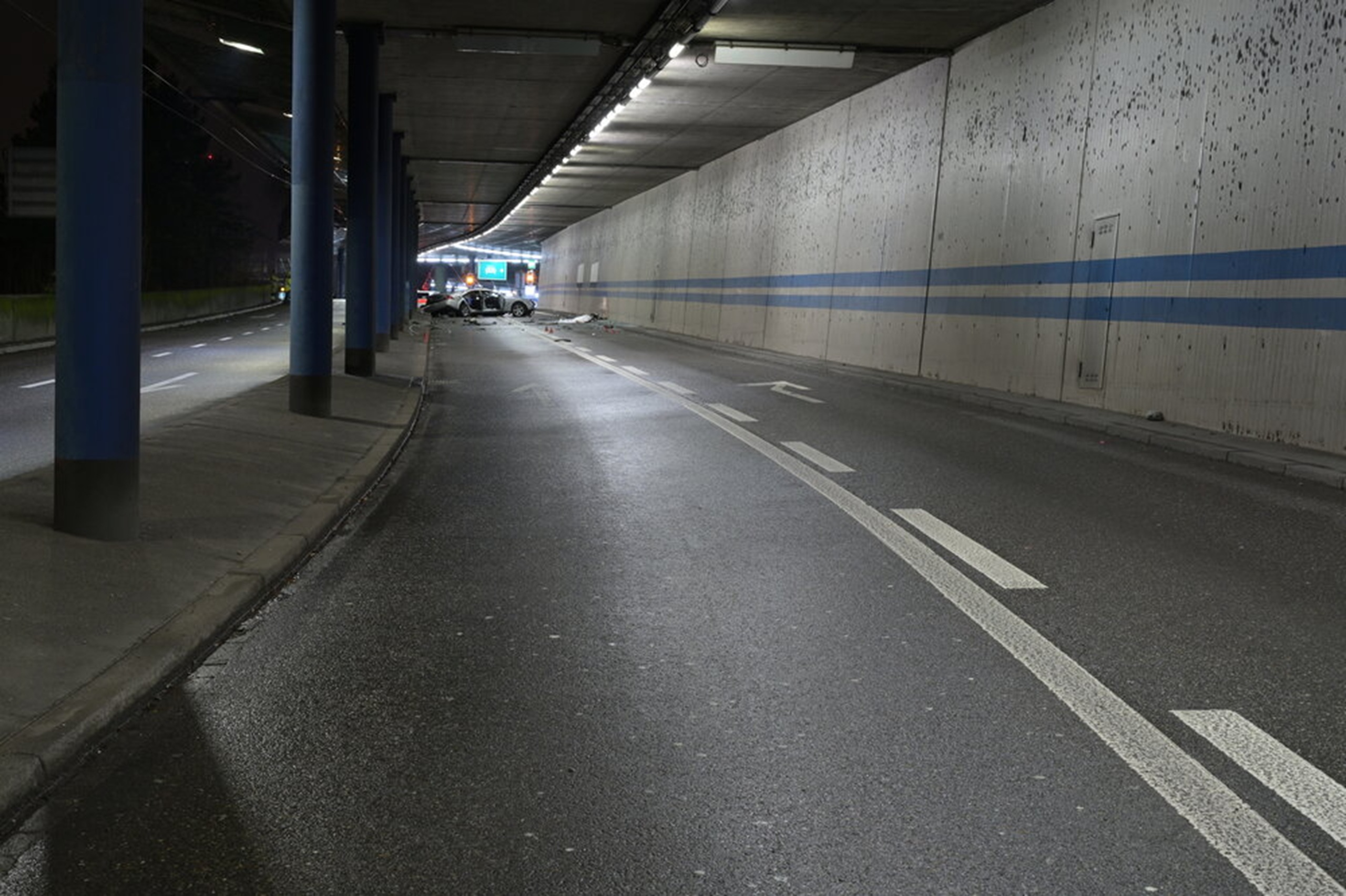 Übersichtsaufnahme einer nassen, tunnelartigen Strasse bei Nacht; in der Ferne steht ein stark beschädigtes Auto, Trümmer liegen auf der Fahrbahn, rechts verläuft eine Betonwand mit blauen Streifen.